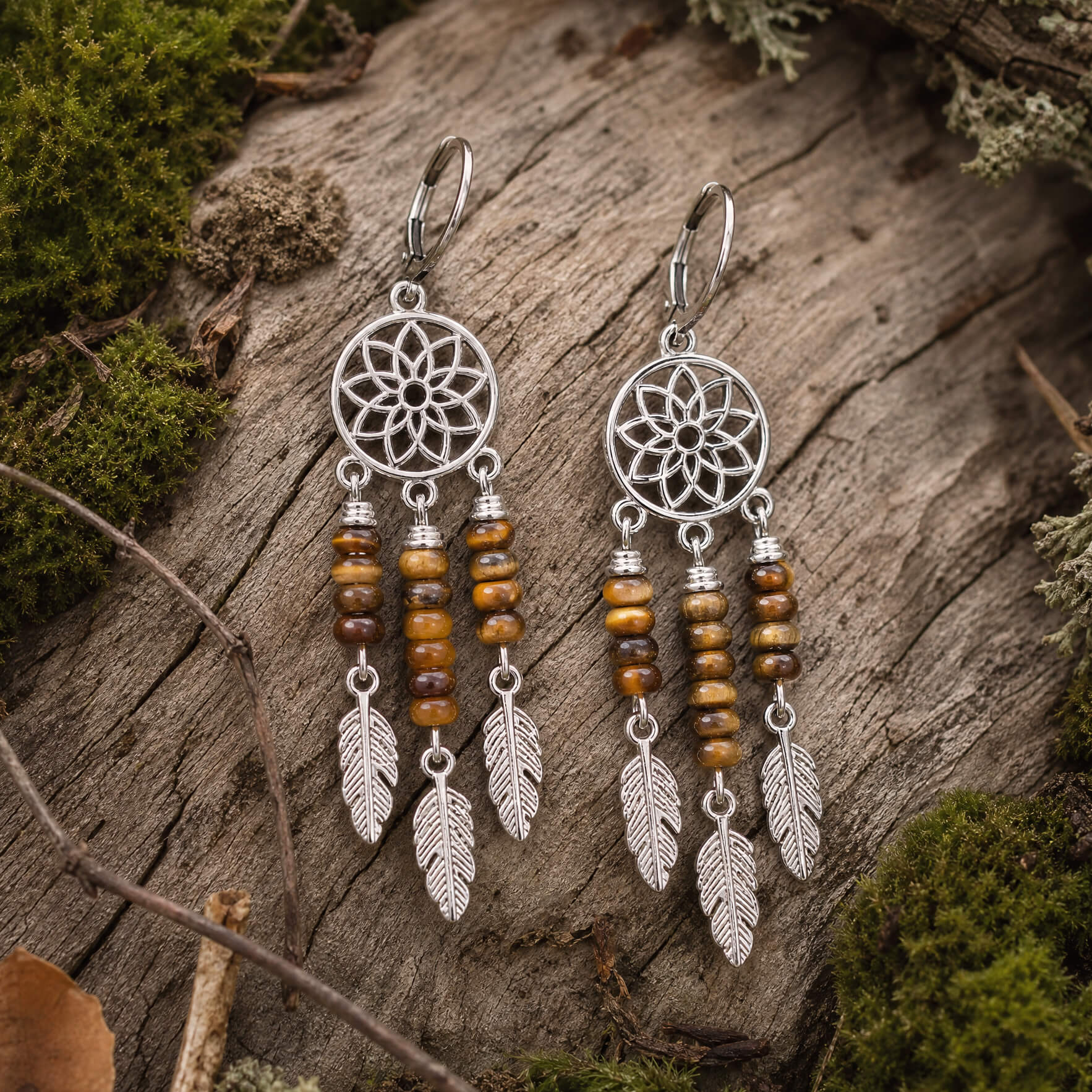 Pair of silver dreamcatcher earrings with tiger's eye beads and feathers on a wooden surface.