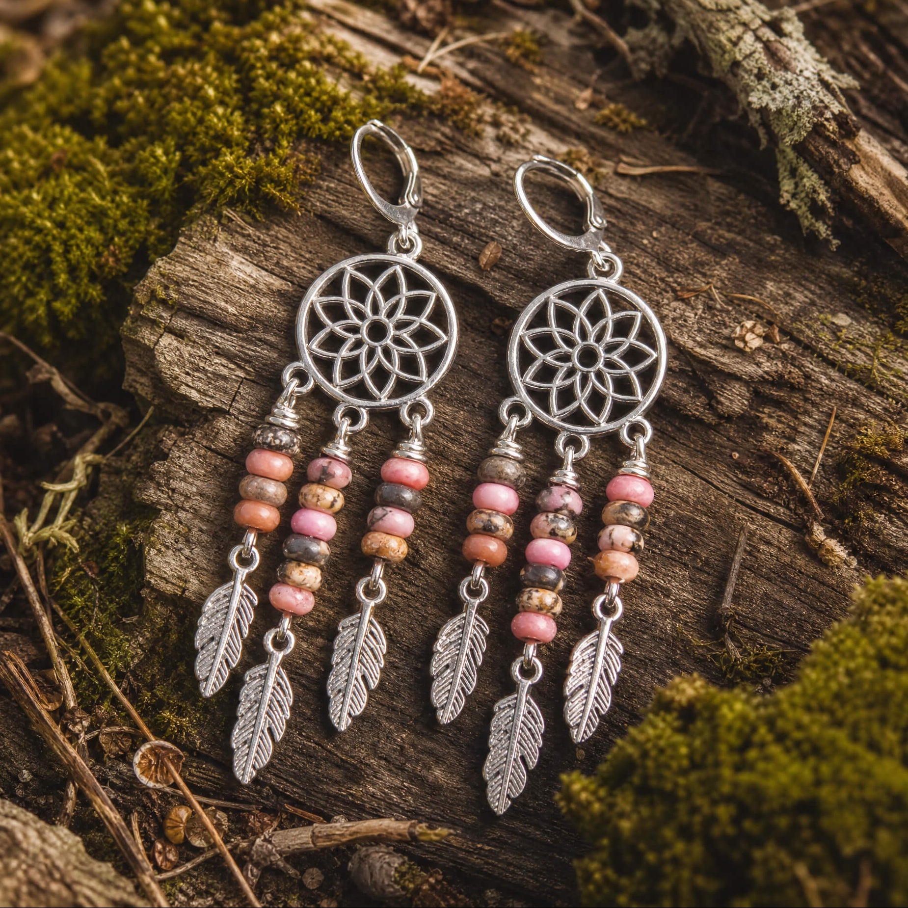 Pair of dreamcatcher earrings with rhodonite beads and feathers on a wooden surface with moss.